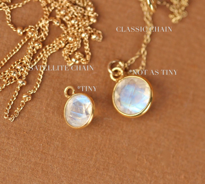 May include: Two gold necklaces with moonstone pendants. The necklace on the left has a satellite chain and a smaller pendant labeled "*TINY". The necklace on the right has a classic chain and a larger pendant labeled "*NOT AS TINY".