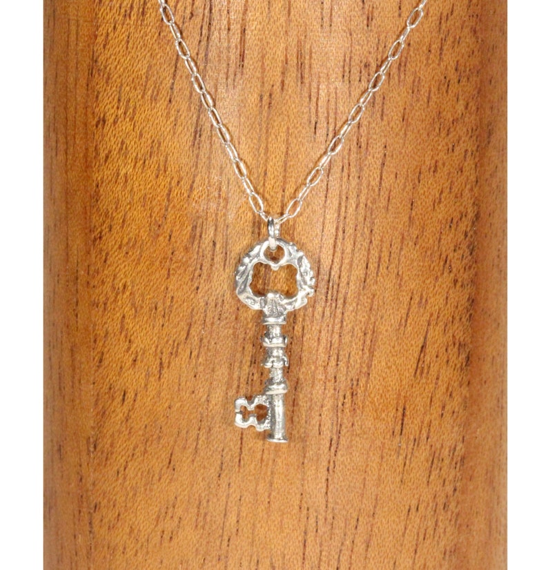 Puede incluir: Un collar con un colgante de llave de plata con un dise&ntilde;o ornamentado. La llave tiene una abertura en forma de coraz&oacute;n y un tallo detallado. El collar est&aacute; en una cadena de plata.