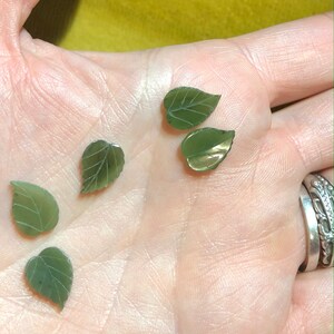 May include: Five green jade leaf-shaped pendants, carved with detailed veins, are displayed on a hand. The pendants are a light green color with a slightly darker green hue in the center.