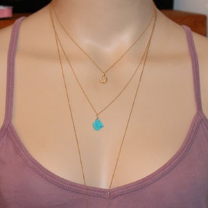May include: A layered gold necklace with three pendants. The top pendant is a small, round, white stone. The bottom pendant is a larger, turquoise stone.