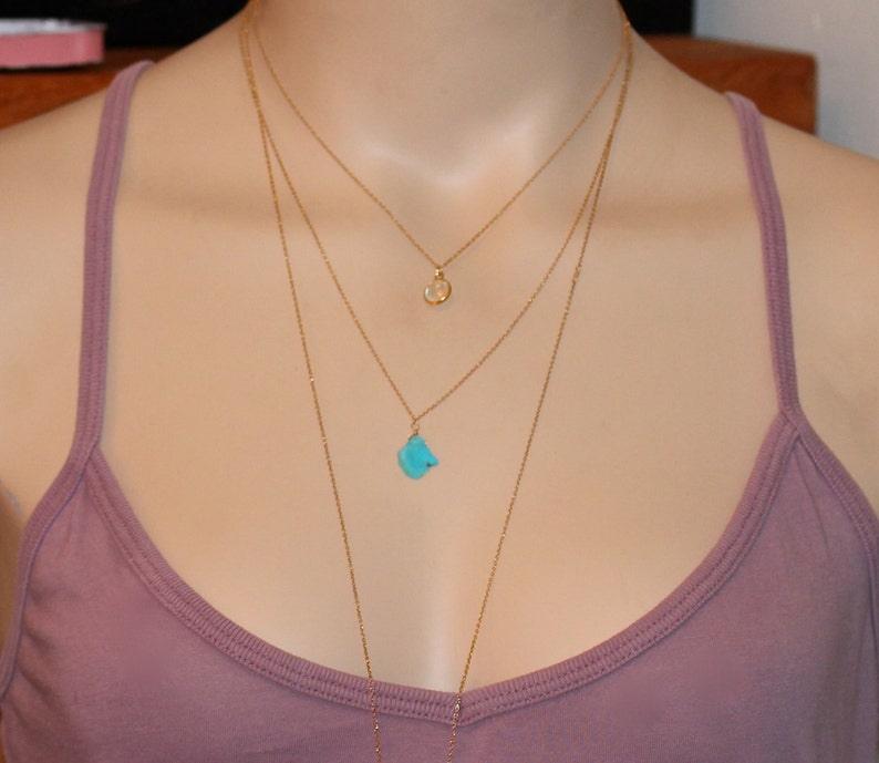 May include: A layered gold necklace with three pendants. The top pendant is a small, round, white stone. The bottom pendant is a larger, turquoise stone.