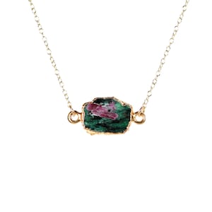 May include: A gold-colored chain necklace featuring a raw, uncut gemstone pendant. The pendant is a mix of green and pink hues, with a gold-colored trim. The necklace is displayed against a white background.