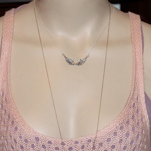 Puede incluir: Un collar de plata con un delicado colgante de ala. El collar est&aacute; superpuesto a una camiseta de tirantes de punto rosa.