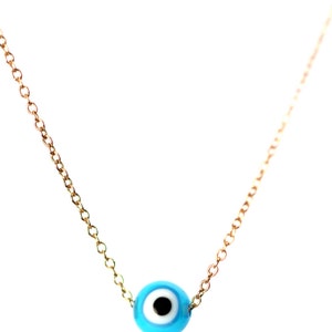 Puede incluir: Un collar de cadena dorada con un dije azul de ojo turco. El dije tiene un centro blanco y una pupila negra.