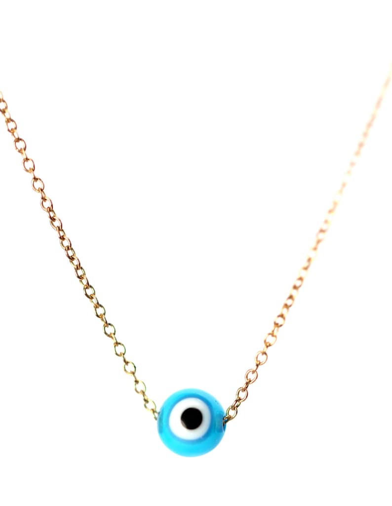 Puede incluir: Un collar de cadena dorada con un dije azul de ojo turco. El dije tiene un centro blanco y una pupila negra.