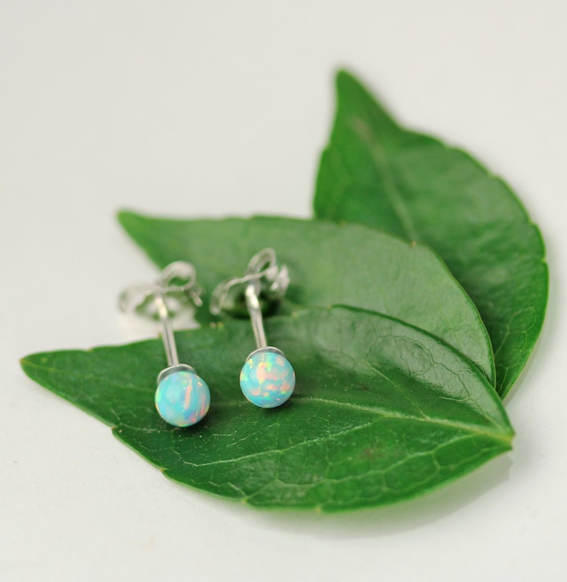 May include: A pair of silver stud earrings with round, iridescent opal stones. The stones are a light blue color with hints of pink and white.