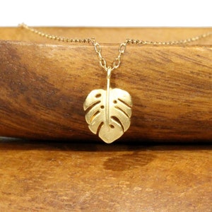 Peut inclure: Un collier en or avec un pendentif d&eacute;licat en forme de feuille de monstera. Le collier est pos&eacute; sur une surface en bois.