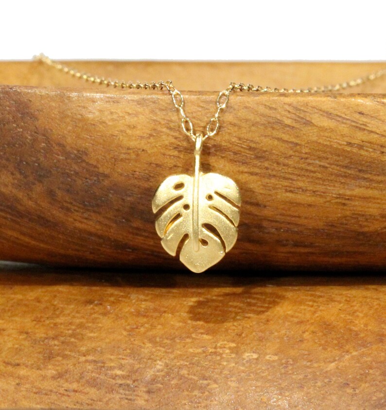 Peut inclure: Un collier en or avec un pendentif d&eacute;licat en forme de feuille de monstera. Le collier est pos&eacute; sur une surface en bois.