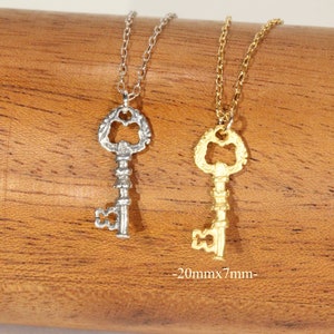 Puede incluir: Dos collares con forma de llave, uno plateado y otro dorado, colgando de una superficie de madera. Los colgantes con forma de llave son ornamentados y miden 20 mm x 7 mm.