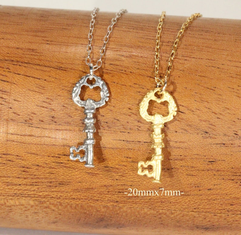 Puede incluir: Dos collares con forma de llave, uno plateado y otro dorado, colgando de una superficie de madera. Los colgantes con forma de llave son ornamentados y miden 20 mm x 7 mm.
