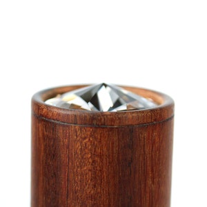 May include: A cylindrical wooden object with a rich brown grain, featuring a clear, faceted, diamond-shaped crystal set into the top opening. The wood has a smooth, polished finish, and the crystal reflects light with sharp, geometric edges.