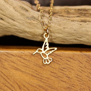 May include: A gold chain necklace with a delicate hummingbird pendant. The hummingbird is cut out of metal and has a detailed design.