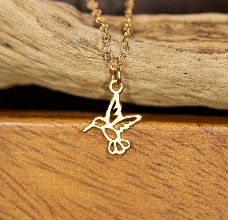 May include: A gold chain necklace with a delicate hummingbird pendant. The hummingbird is cut out of metal and has a detailed design.