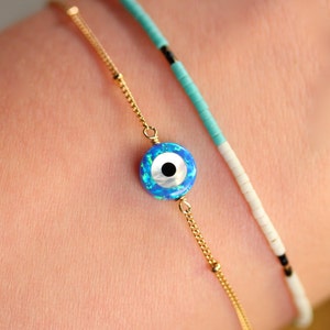 May include: A gold chain bracelet with a blue and white evil eye charm. The bracelet is layered with a turquoise and white beaded bracelet.