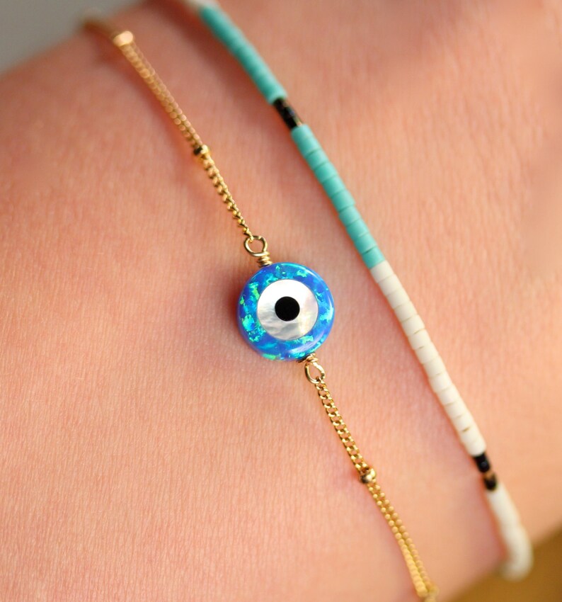 May include: A gold chain bracelet with a blue and white evil eye charm. The bracelet is layered with a turquoise and white beaded bracelet.