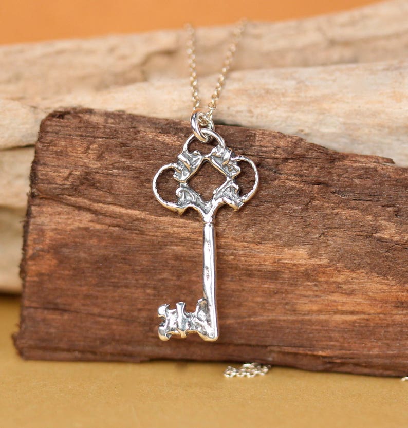 May include: Silver key pendant necklace with an ornate design. The key has a detailed, intricate design with a heart-shaped top and a long, slender shaft.