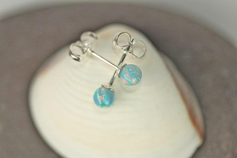 May include: A pair of silver stud earrings with blue opal stones. The earrings are displayed on a white seashell.