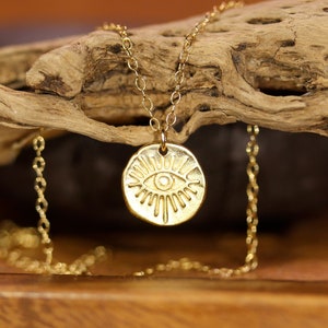 May include: Gold necklace with a round pendant featuring a stylized eye design. The pendant has a textured surface and is surrounded by radiating lines.