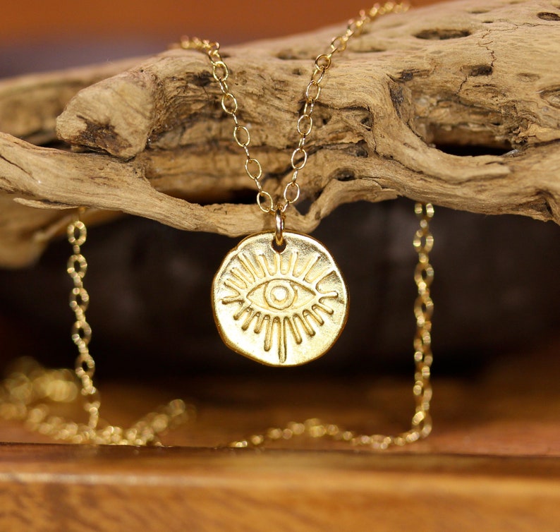 May include: Gold necklace with a round pendant featuring a stylized eye design. The pendant has a textured surface and is surrounded by radiating lines.