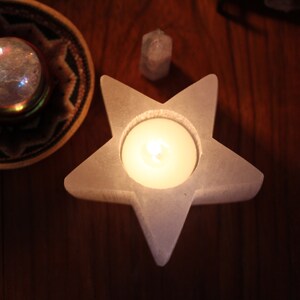 May include: A white star-shaped candle holder with a lit candle inside. The holder is made of a smooth, white material and has a slightly textured surface. The candle is a small, round, white candle.