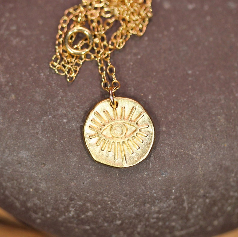 May include: Gold necklace with a round pendant featuring a stylized eye design. The pendant has a textured surface and a raised eye with radiating lines.