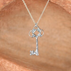May include: A silver key pendant necklace with an ornate design. The key has a detailed, intricate design with a decorative top and a textured body.