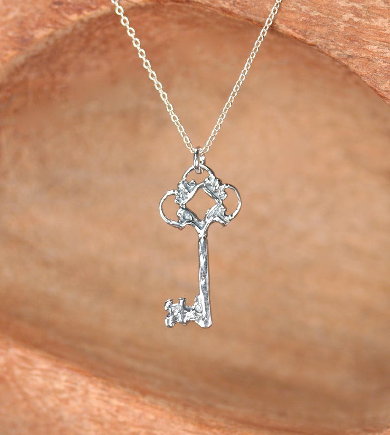 May include: A silver key pendant necklace with an ornate design. The key has a detailed, intricate design with a decorative top and a textured body.