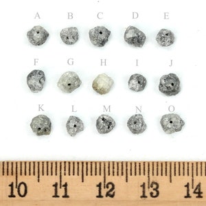 May include: Fifteen rough, grey, diamond beads with holes drilled through the centre. The beads are arranged in three rows of five, labelled A through O. A ruler is shown below the beads, with markings from 10 to 14 inches.