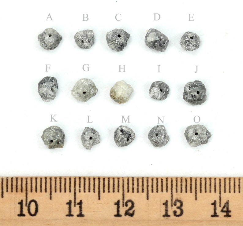 May include: Fifteen rough, grey, diamond beads with holes drilled through the centre. The beads are arranged in three rows of five, labelled A through O. A ruler is shown below the beads, with markings from 10 to 14 inches.