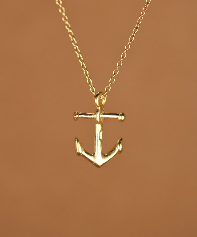May include: A gold anchor pendant necklace. The anchor is a simple design with a rounded top and a slightly curved bottom.