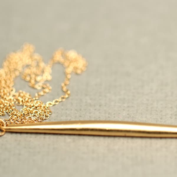 May include: A gold chain necklace with a long, thin, gold pendant. The pendant is shaped like a long, thin, pointed blade.