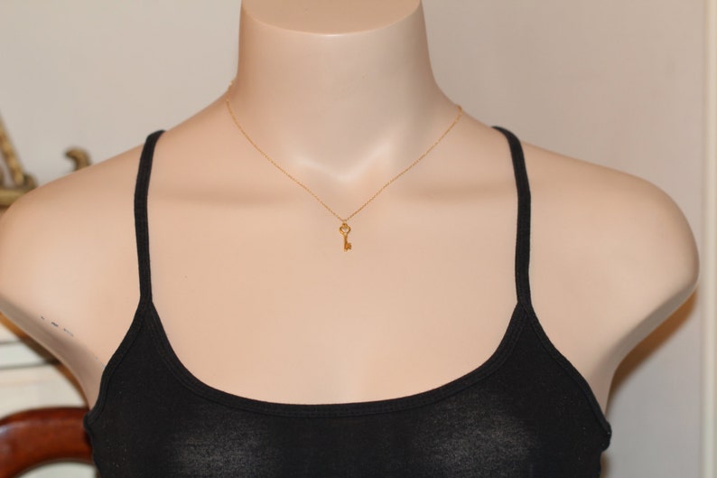 Puede incluir: Un collar con un colgante de llave de oro en una cadena de oro. La llave es un dise&ntilde;o simple con un lazo en forma de coraz&oacute;n en la parte superior.