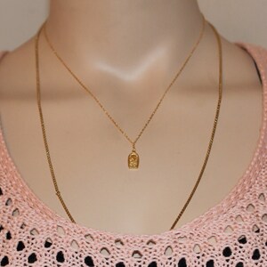 May include: A gold chain necklace with a small pendant featuring a detailed design. The necklace is layered over a second, thinner gold chain.
