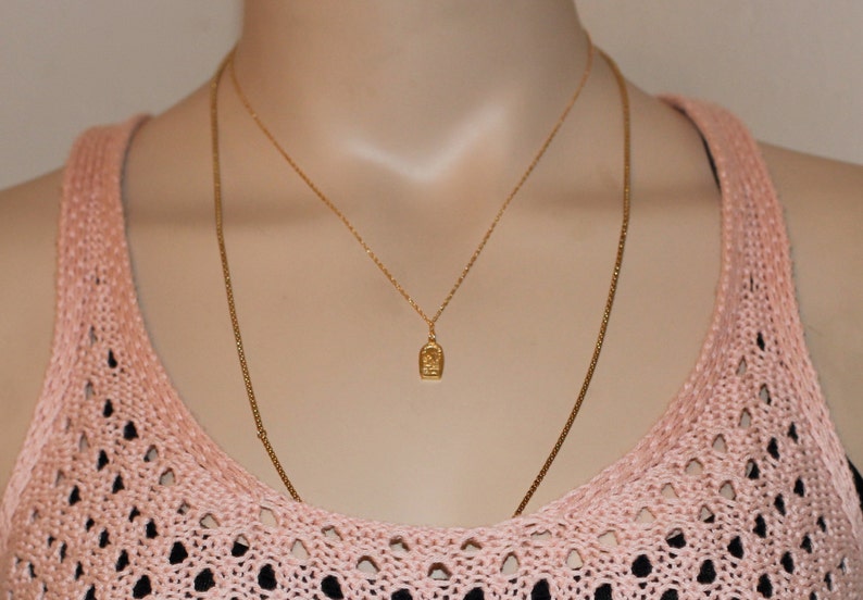 May include: A gold chain necklace with a small pendant featuring a detailed design. The necklace is layered over a second, thinner gold chain.