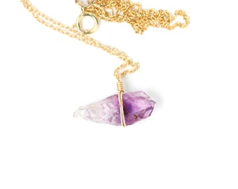 May include: A gold chain necklace with a raw amethyst crystal pendant. The crystal is wrapped in gold wire and has a purple and clear color.