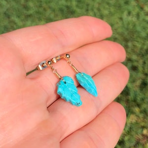 May include: A pair of turquoise leaf earrings with gold wire and posts. The earrings are held in a hand.