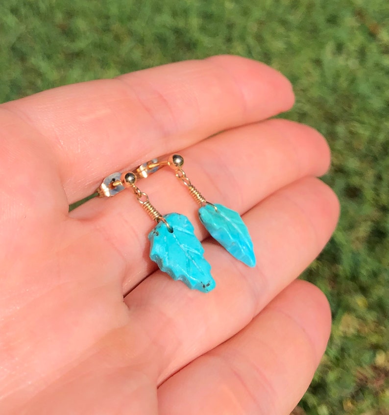 May include: A pair of turquoise leaf earrings with gold wire and posts. The earrings are held in a hand.