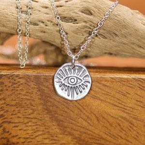 May include: Silver necklace with a round pendant featuring a stylized eye with radiating lines. The necklace is on a wooden surface.