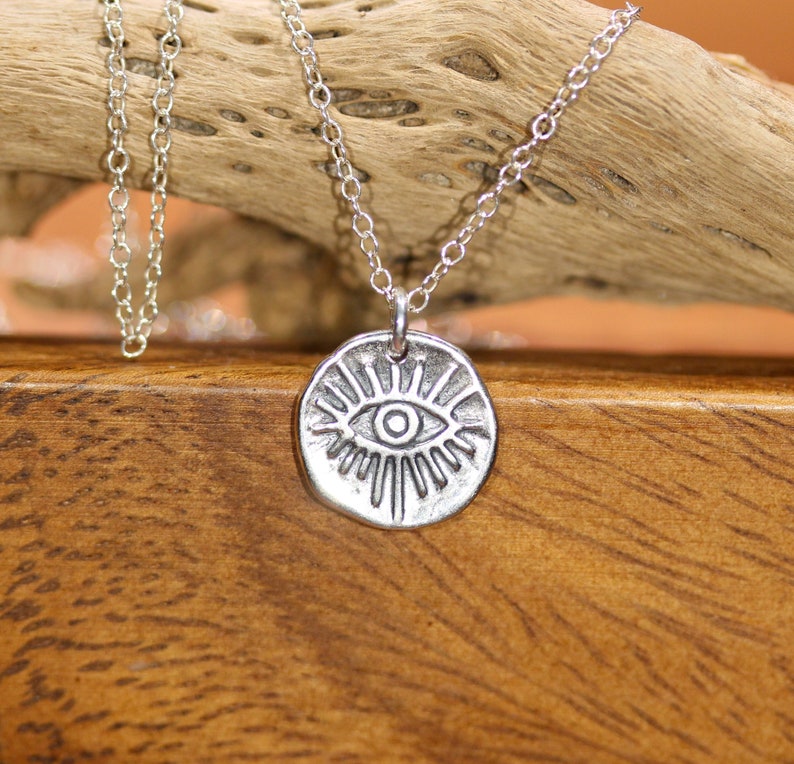 May include: Silver necklace with a round pendant featuring a stylized eye with radiating lines. The necklace is on a wooden surface.