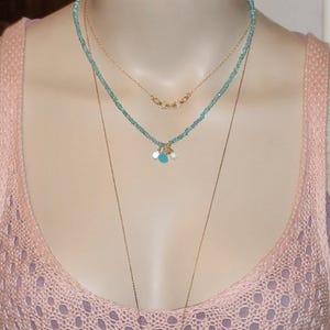 May include: A layered necklace with a delicate gold chain and a turquoise beaded chain with small gold charms. The necklace is worn by a person with a pink knitted top.