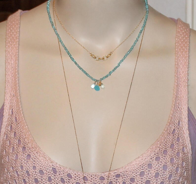 May include: A layered necklace with a delicate gold chain and a turquoise beaded chain with small gold charms. The necklace is worn by a person with a pink knitted top.