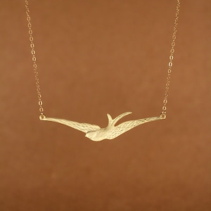 Puede incluir: Un collar de oro con una cadena delicada y un colgante con forma de golondrina volando.