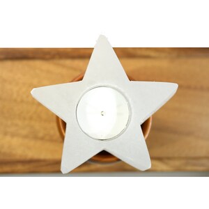 May include: A white ceramic star-shaped candle holder with a round opening for a tea light. The holder is sitting on a wooden surface.