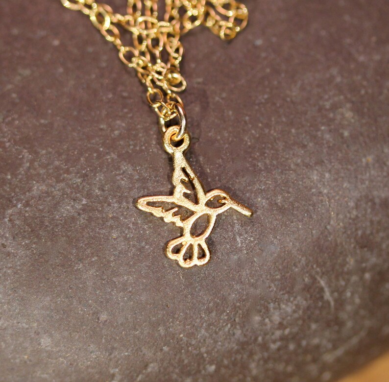 May include: A gold chain necklace with a delicate hummingbird pendant. The hummingbird is cut out of metal and has a detailed design.