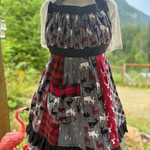 May include: A black and red patchwork apron with a deer print and a large pocket. The apron has a black ruffle around the bottom.
