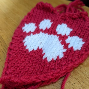 May include: A red knitted heart with a white paw print design. The heart is made of yarn and has a textured surface.