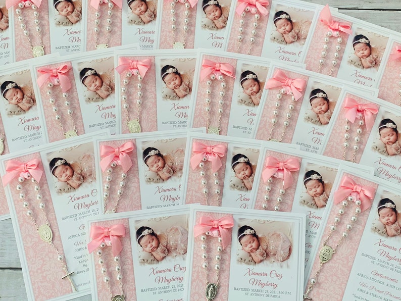 May include: Pink and white baptism favors with a photo of a baby girl, a silver rosary, and a pink ribbon. The card reads "Xiamara Cruz Mayberry Baptized March 28, 2021, 1:00 PM St. Anthony de Padua"