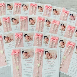May include: Pink and white baptism favors with a pearl rosary, a small silver cross, and a photo of a baby girl. The card includes the baby's name, date of baptism, and a religious quote.