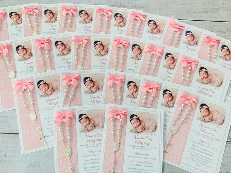 May include: Pink and white baptism favors with a pearl rosary, a small silver cross, and a photo of a baby girl. The card includes the baby's name, date of baptism, and a religious quote.