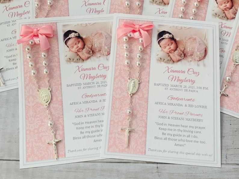 May include: A pink and white card with a silver rosary attached. The card has a photo of a baby girl and text that reads "Xiamara Cruz Mayberry Baptized March 28, 2021, 3:00 PM St. Anthony de Padua Godparents Africa Miranda & Jed Lowrie Her Proud Parents John & Stefani Mayberry "God in Heaven hear my prayer. Keep me in thy loving care. Be my guide in all I do. Bless all those who love me too. Amen" Thank you for sharing this special day with us!"